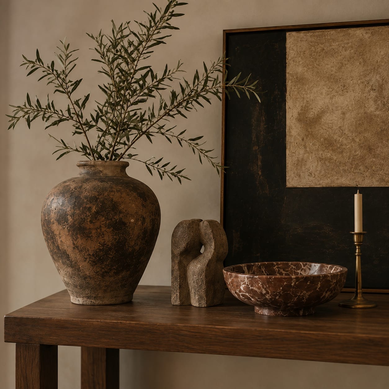 Objects arranged along a dark oak console against a plastered wall: an aged terracotta vessel holding olive branches, a small carved stone sculpture pierced through its centre, a shallow bowl in oxblood-and-white marble, and a slim brass candlestick with a pale unlit candle. A large dark canvas in a timber frame leans on the wall behind them.